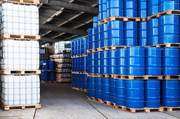 Blue steel drums stacked on pallets beside IBC totes in a chemical storage warehouse